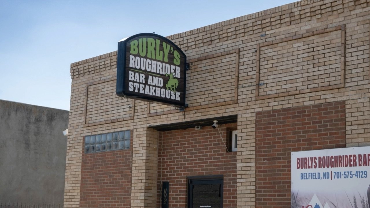 The brick storefront of Burly's Roughrider on Main Street in Belfield, North Dakota, with the lit Bar and Steakhouse sign projecting over the door